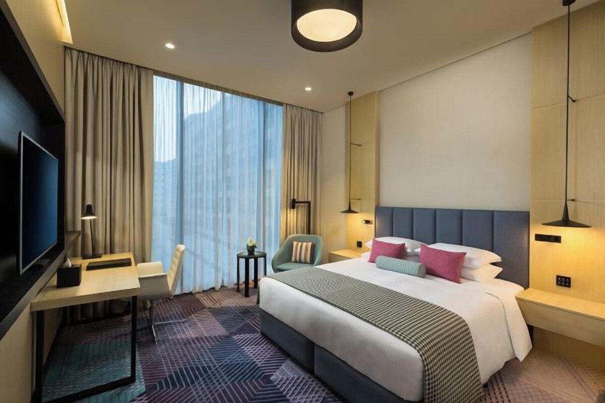 A modern hotel room with a large bed, a desk, a TV, and a large window with curtains.