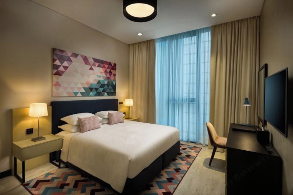 A hotel room with a large bed, two bedside tables with lamps, a desk with a chair, a TV, and a geometric-patterned rug.