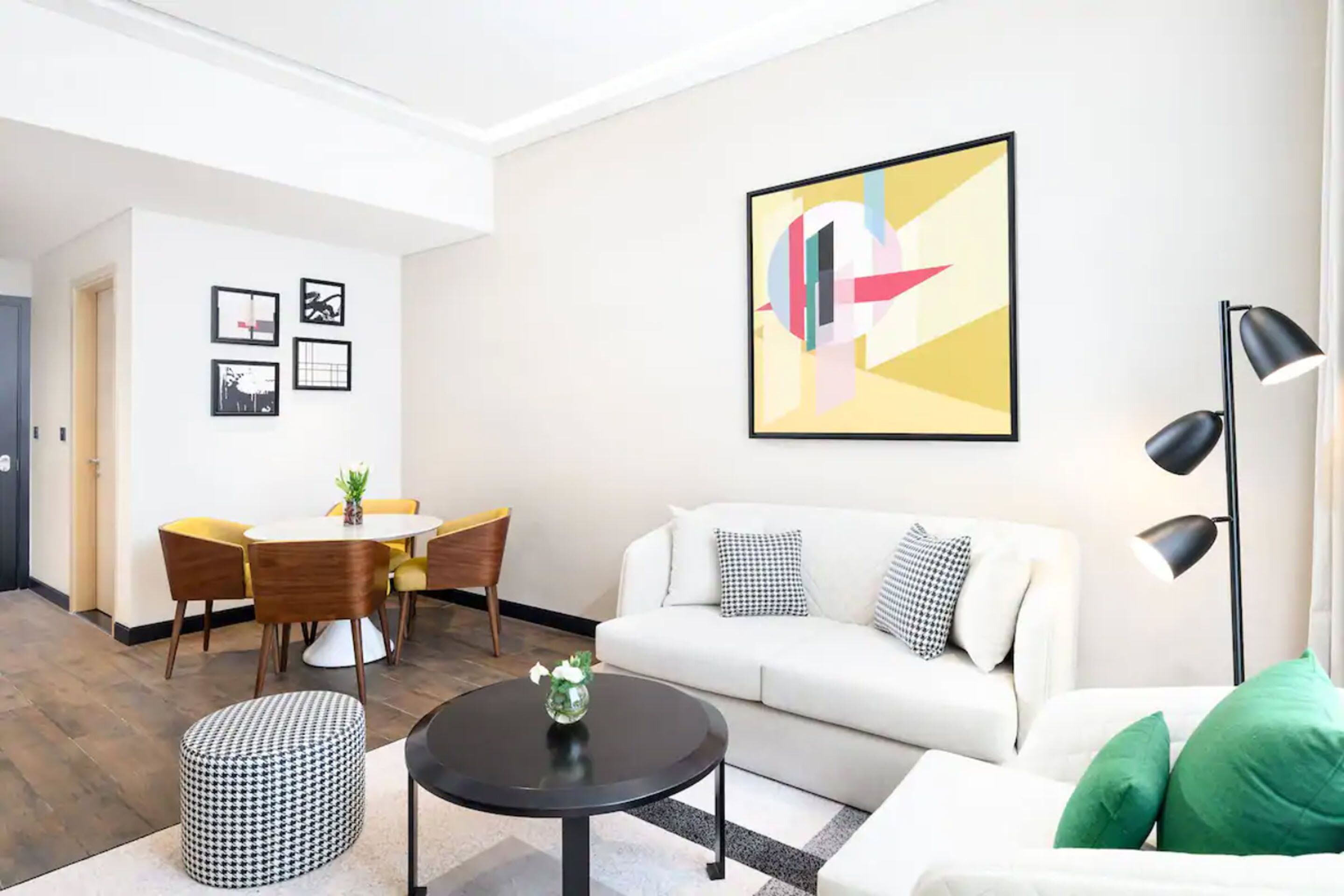 A modern living room with a white sofa, a round black coffee table, a houndstooth ottoman, a dining area with a round table and chairs, and a large abstract painting on the wall.