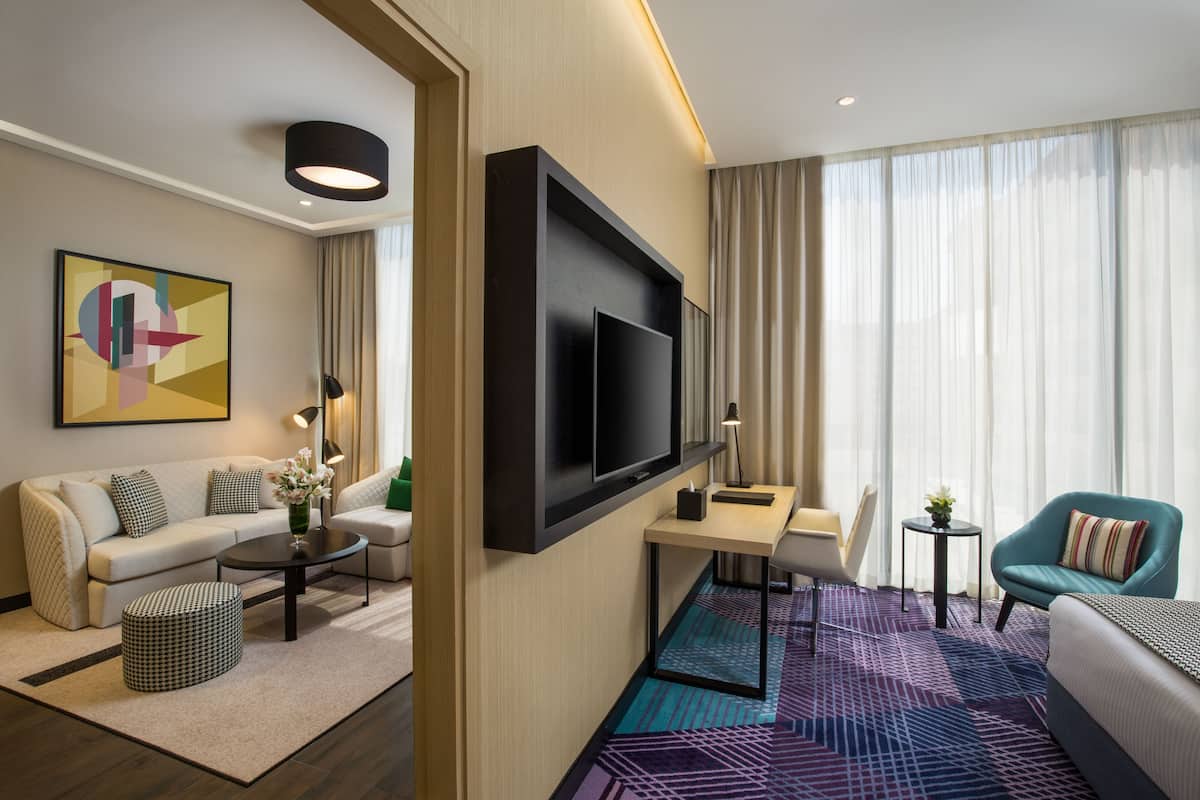 Moderni hotellihuone, jossa on taulutelevisio, sohva, tuoli, pieni pöytä ja kuviollinen matto.