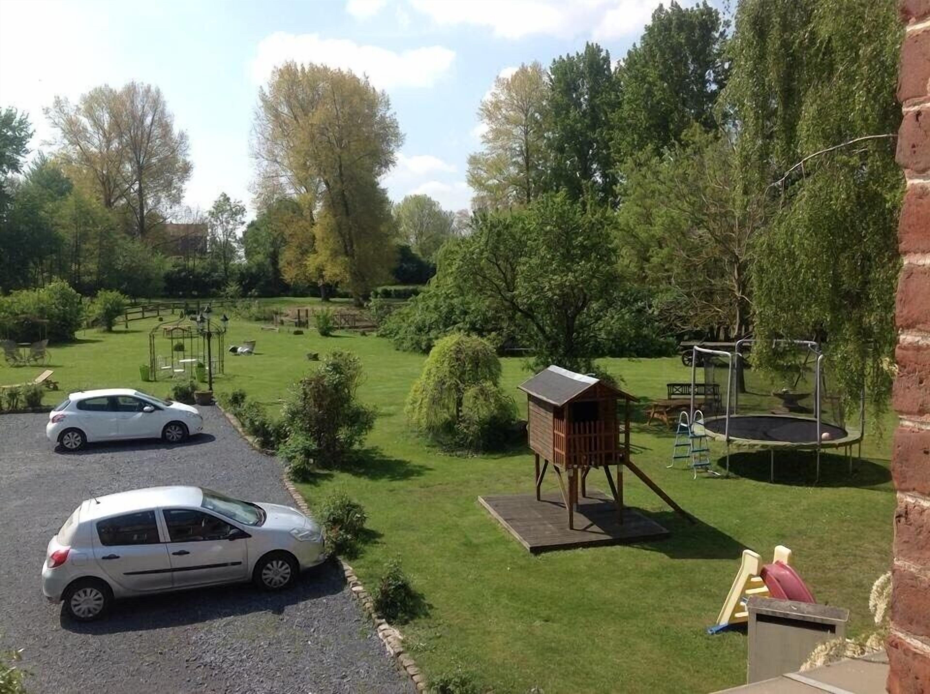Un jardin avec une cabane en bois, un trampoline et une voiture blanche garée au premier plan.