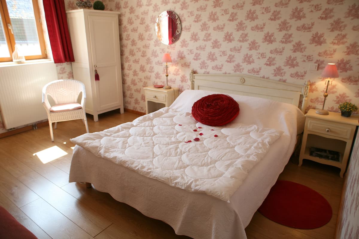 A bedroom with a large bed, a white wardrobe, a bedside table with a lamp, and a chair.