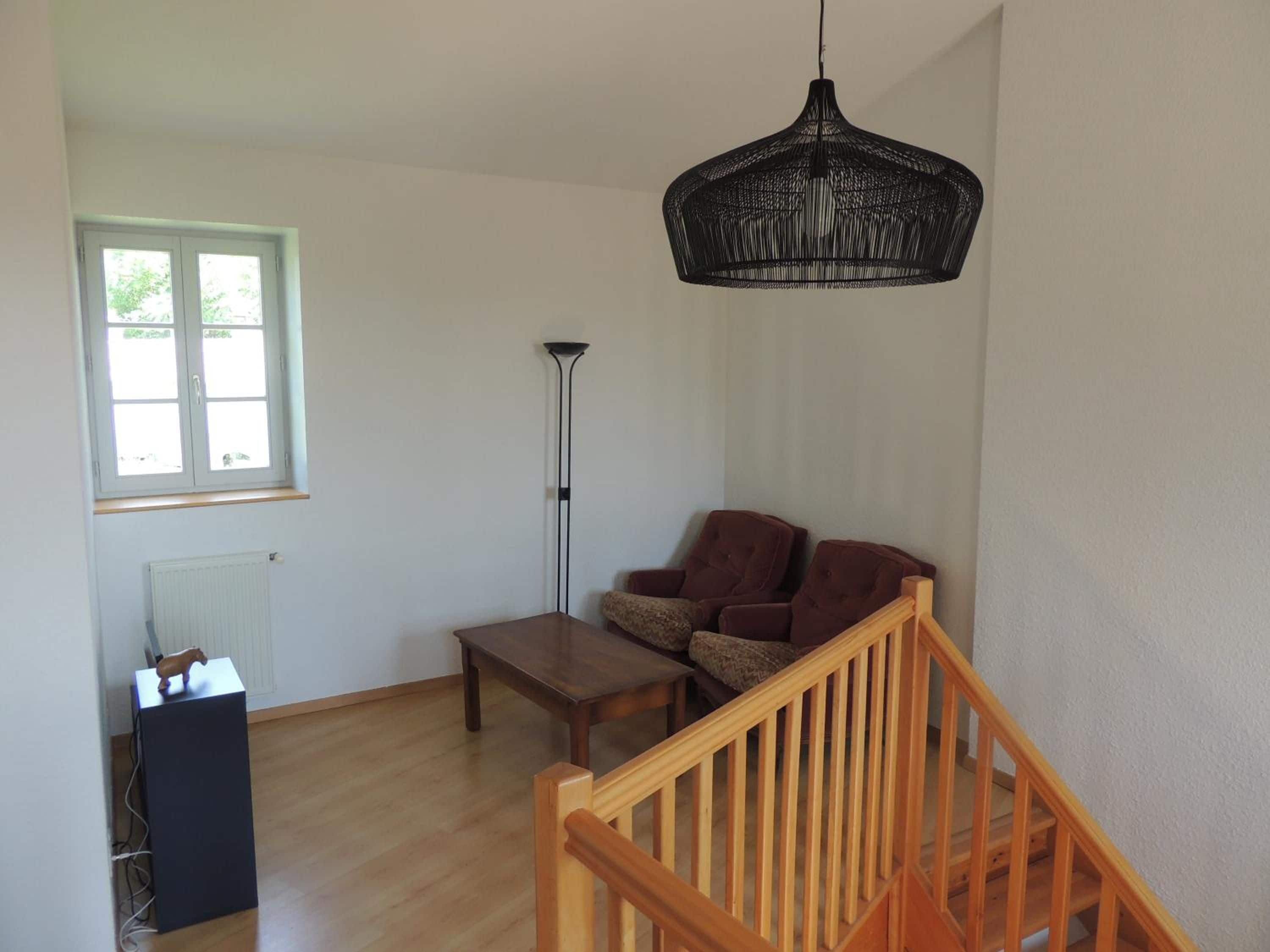 A room with a wooden floor, a sofa, a coffee table, a radiator, a window, and a hanging light fixture.