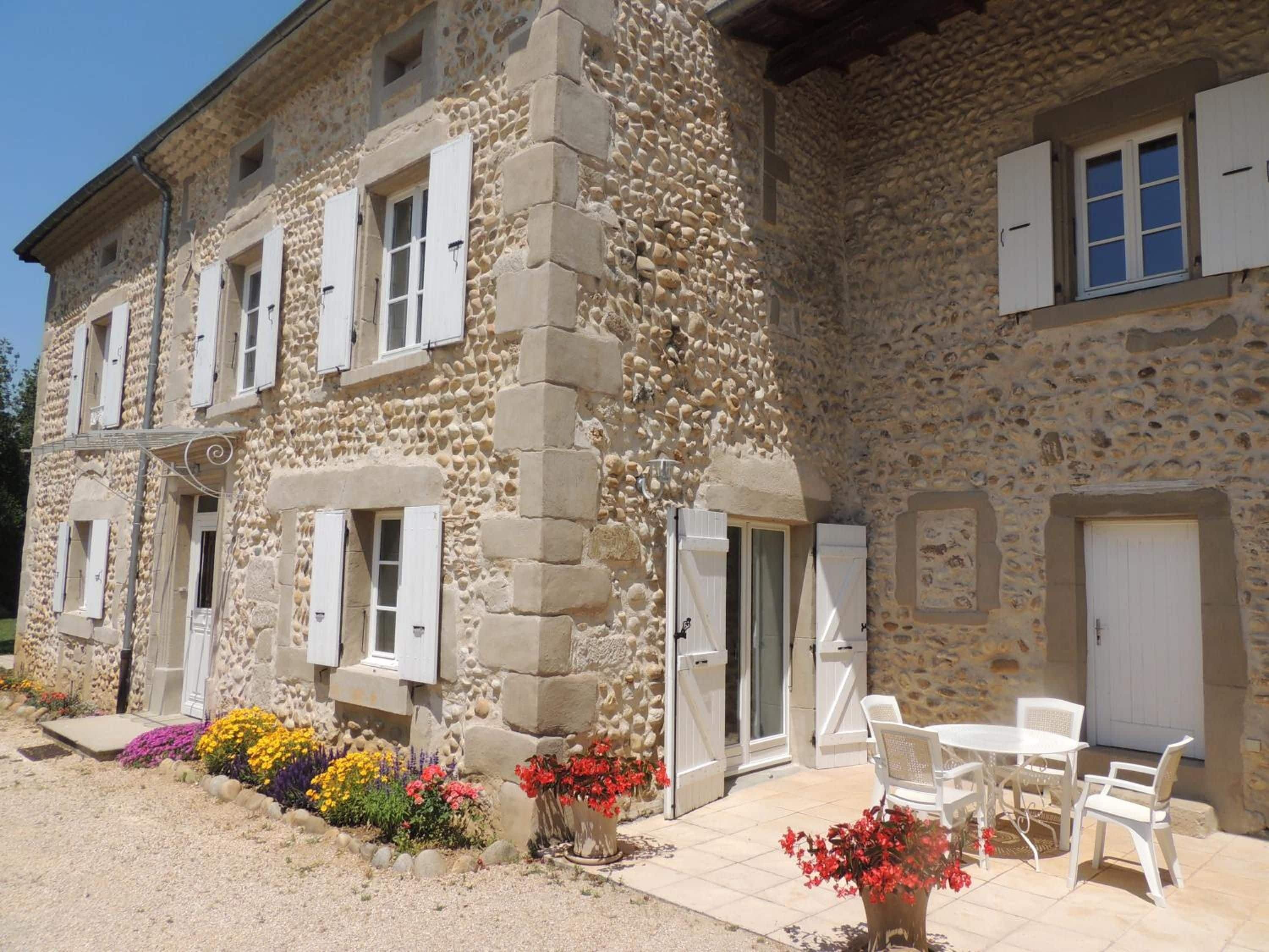 A stone building with multiple windows and doors, outdoor seating area with white chairs and tables, and flower beds with various colorful flowers.