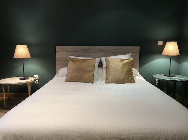 Una cama bien hecha con dos almohadas, una mesita de noche redonda con una lámpara, y otra mesita similar al otro lado de la cama.