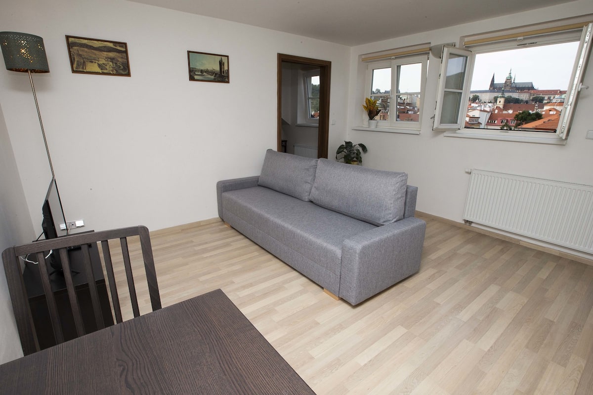 Ein Wohnzimmer mit grauem Sofa, einem Esstisch mit Stühlen und Blick auf eine Stadtlandschaft durch ein Fenster.