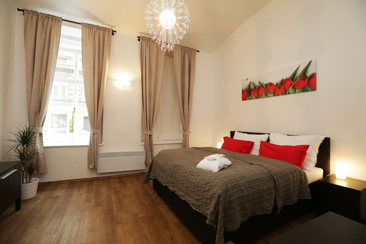 Ein Schlafzimmer mit einem Bett, Vorhängen, einer Chandelüster, einem Gemälde von roten Tulpen und einer Topfpflanze.