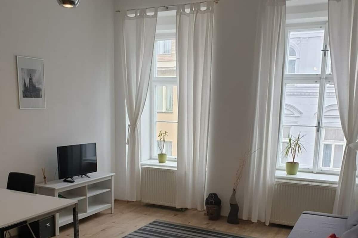Ein modernes Wohnzimmer mit einer Couch, einem Fernseher auf einem Schrank, einem Esstisch und zwei mit Vorhängen versehenen Fenstern.