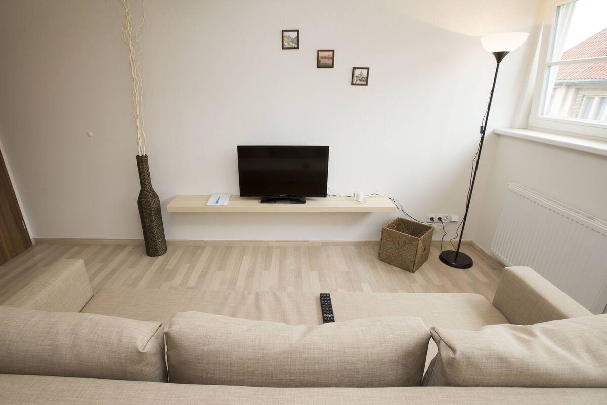 Ein Wohnzimmer mit Flachbildfernseher auf einem Standfuß, einer Couch, einer Stehlampe und einer Vase auf dem Boden.