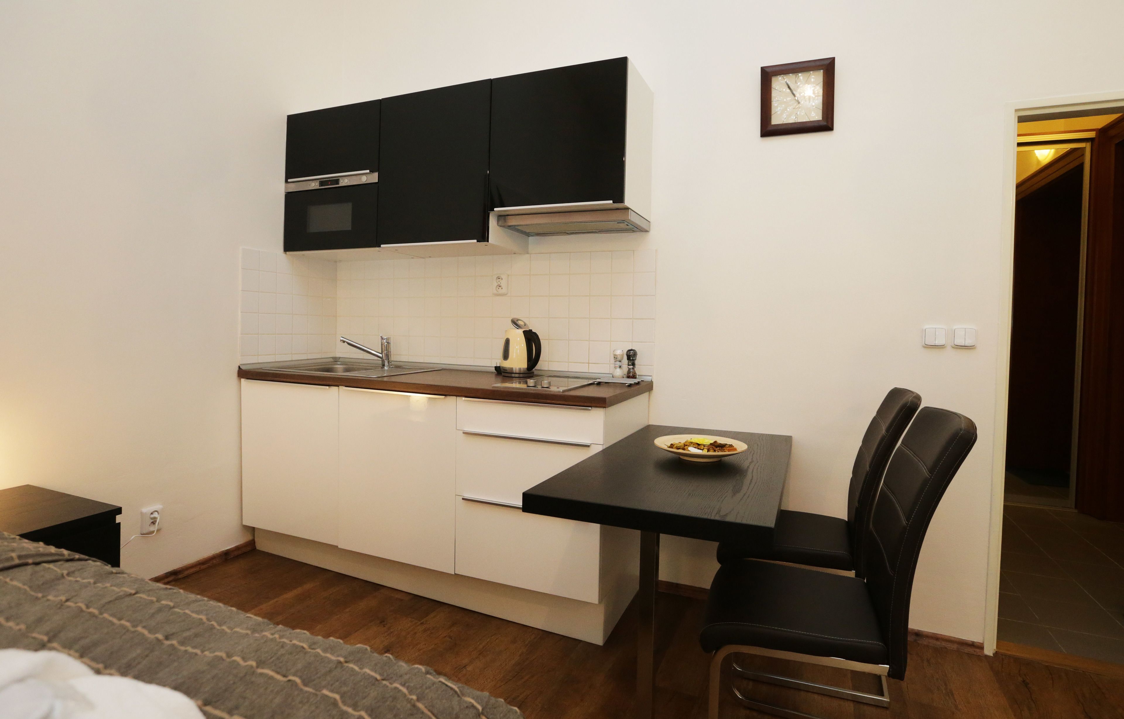 Une cuisine compacte avec des meubles noirs, une cuisinière et une bouilloire posée sur le plan de travail. On y trouve une petite table à manger avec une chaise et un bol de nourriture.
