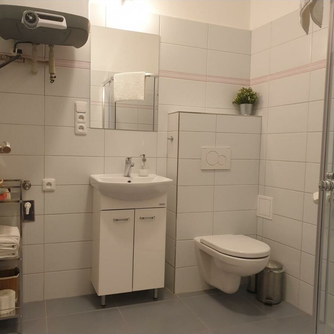 Un baño moderno con lavamanos, espejo y inodoro blancos. Hay una planta pequeña en la pared y un cubo de basura cerca del inodoro.
