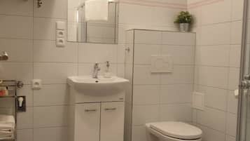 Un baño moderno con lavamanos, espejo y inodoro blancos. Hay una planta pequeña en la pared y un cubo de basura cerca del inodoro.