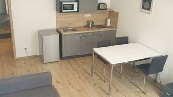 A compact kitchen with a microwave, toaster, and sink. There's a small refrigerator, a dining table with chairs, and a grey sofa.