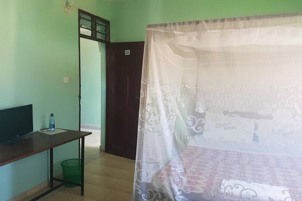 Una habitación con un escritorio, una silla, una botella de agua, una cesta verde y una puerta con un letrero de número.