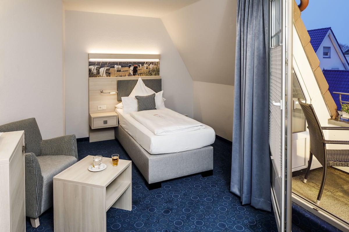 Ein Hotelzimmer mit einem Bett, einem kleinen Tisch mit Glas und Tasse, einem Sessel und Blick ins Freie durch ein Fenster.
