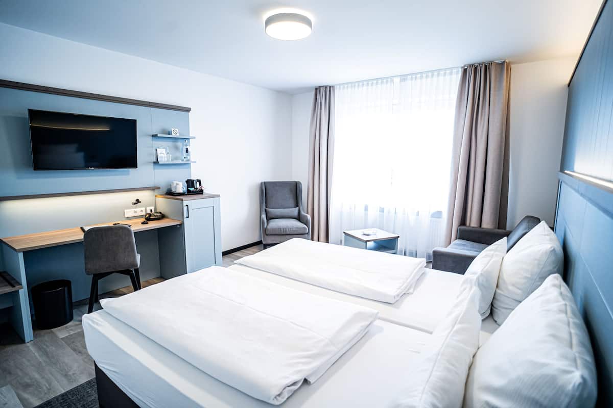 Ein Hotelzimmer mit zwei Betten, einem Schreibtisch, einem Sessel, einem Fernseher und einem Fenster mit Vorhängen.