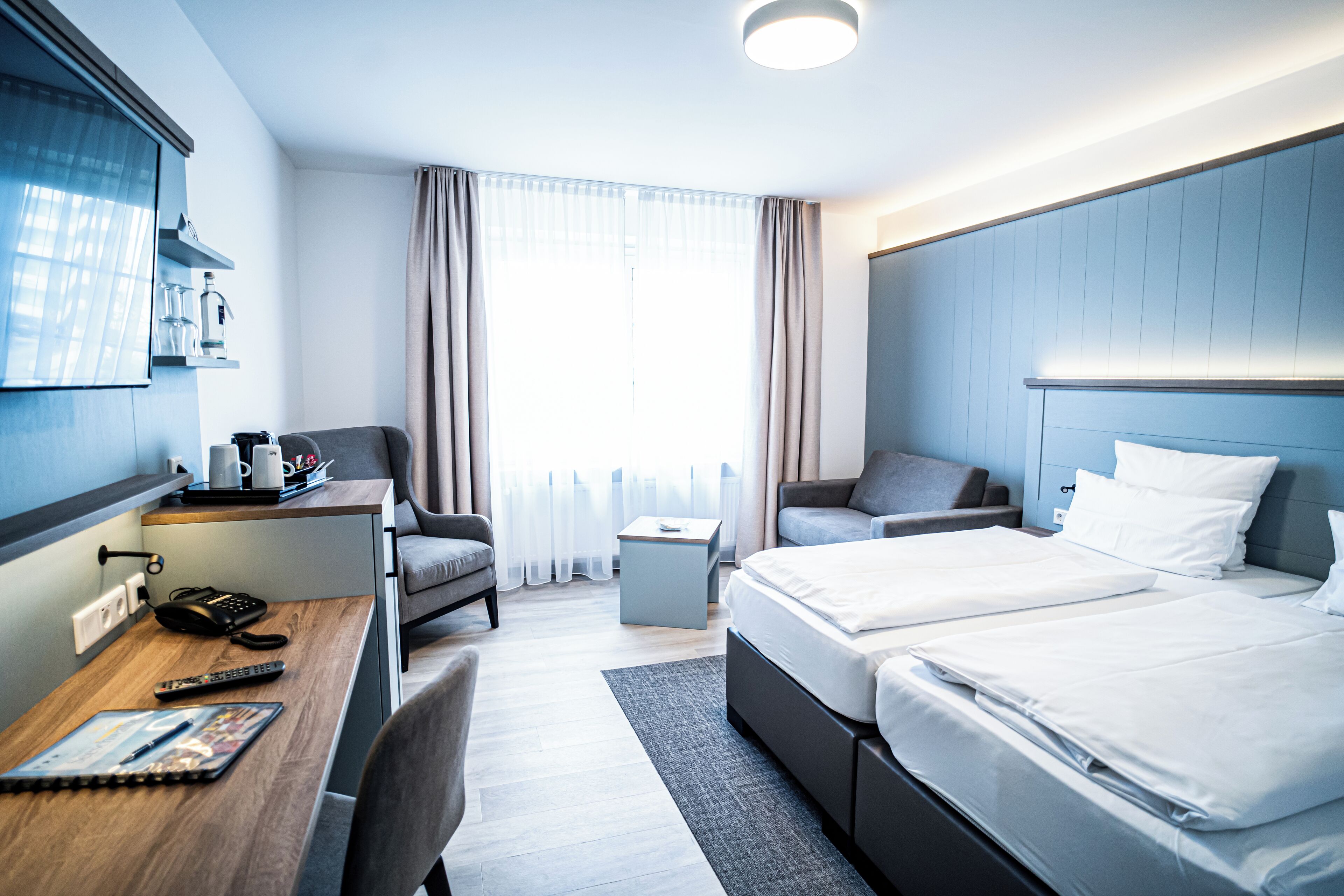 A modern hotel room with a desk, two beds, a sofa, and a small side table.