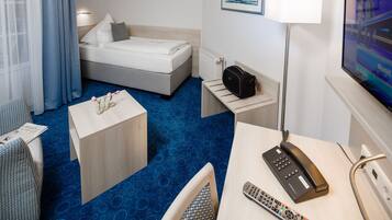 Una habitación de hotel con una cama, un escritorio con teléfono y control remoto, una silla, una mesita con flores, una lámpara y un televisor en la pared.