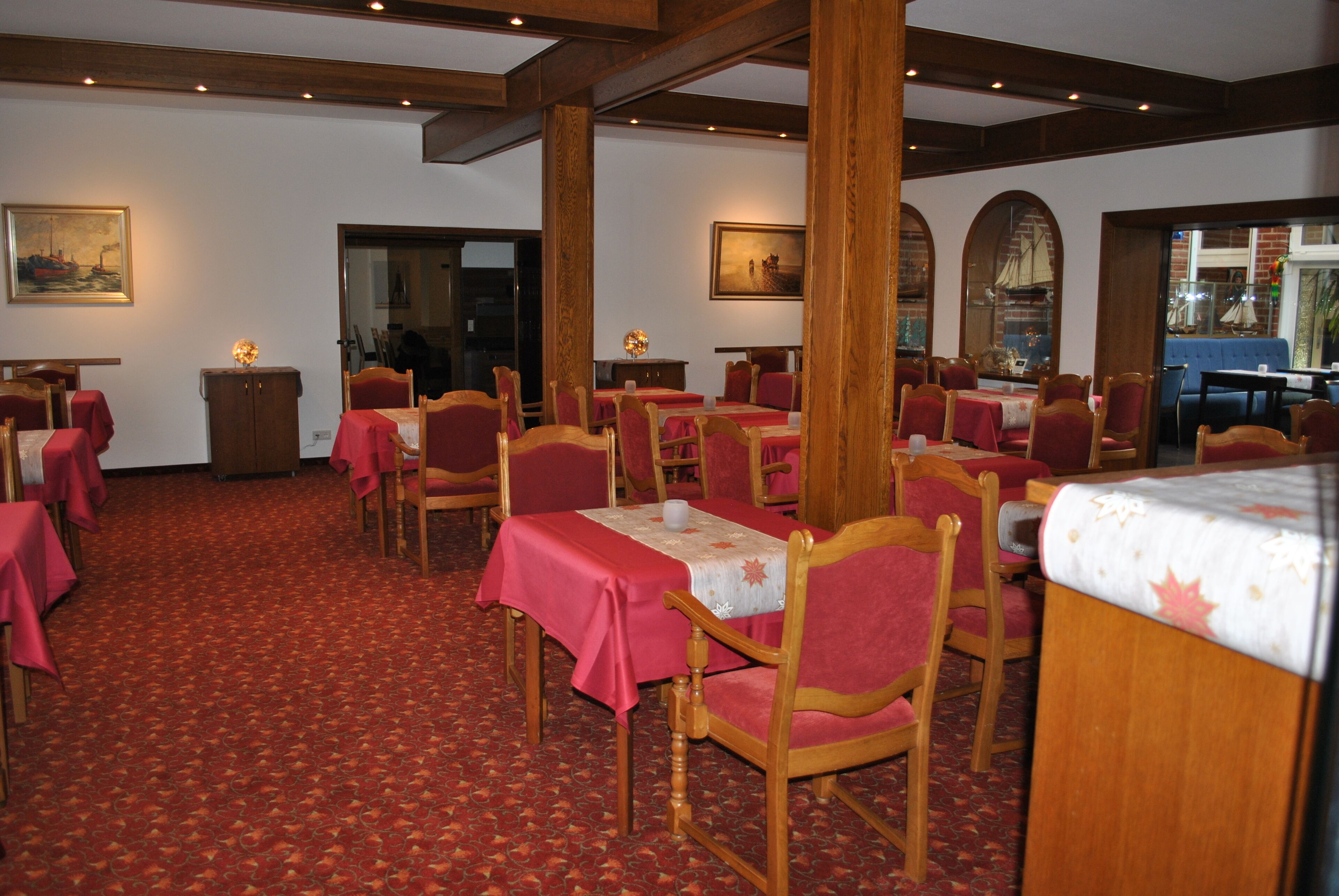 A restaurant with wooden chairs and tables, red carpet flooring, and paintings on the walls.