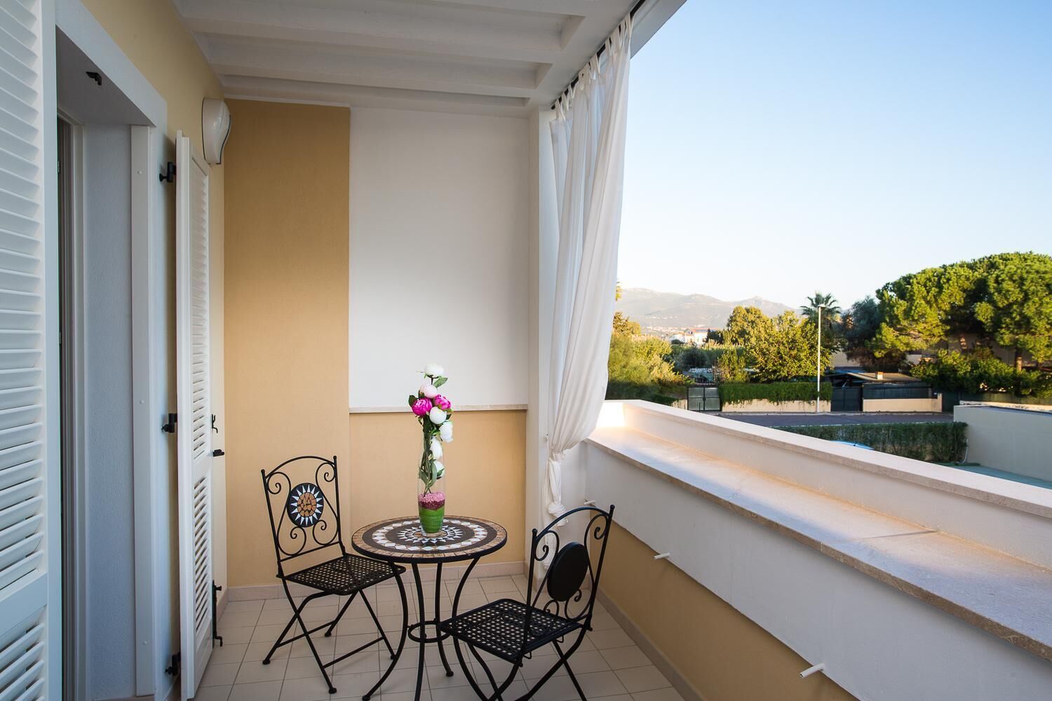 Un balcón con mesa y sillas, un jarrón con flores y vistas a zonas verdes y edificios.