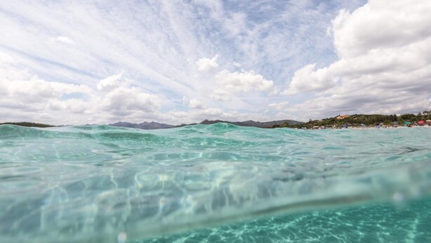 On peut admirer une vue dégagée sur l’océan, dont l’eau est d’un bleu turquoise, avec une côte et des montagnes qui se dessinent à l’horizon.
