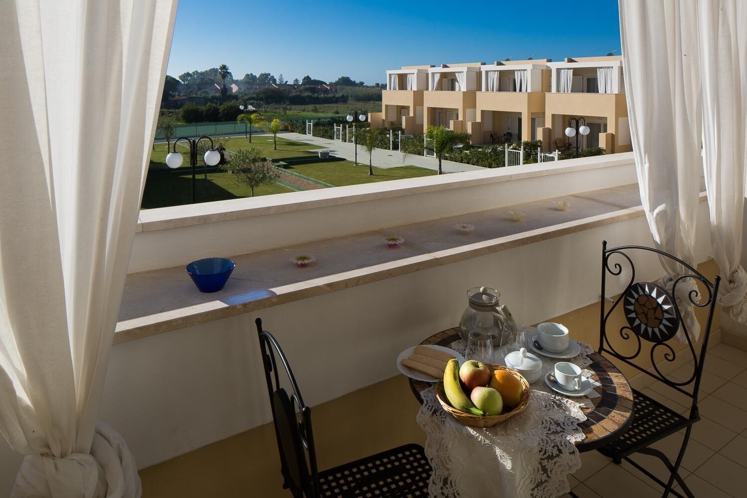 A balcony with a table set for breakfast, a view of a building, and a garden.