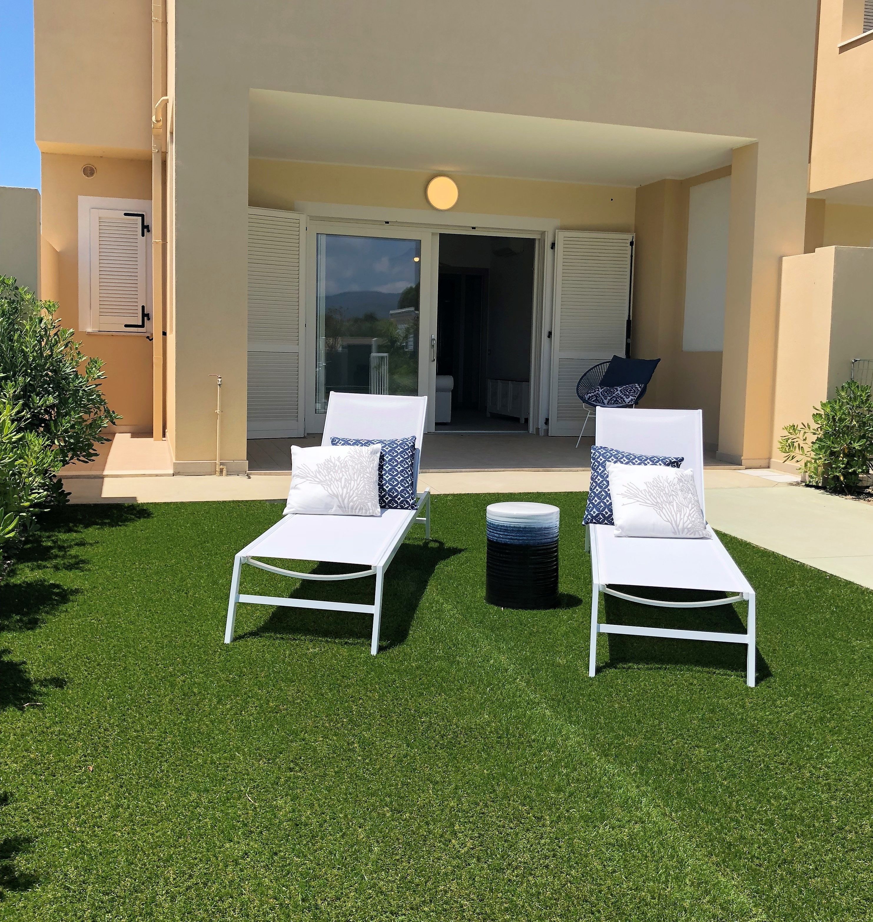 Dos sillones blancos con cojines estampados sobre un césped cuidado, un taburete redondo pequeño y la entrada de un edificio con puerta de vidrio y una lámpara circular.