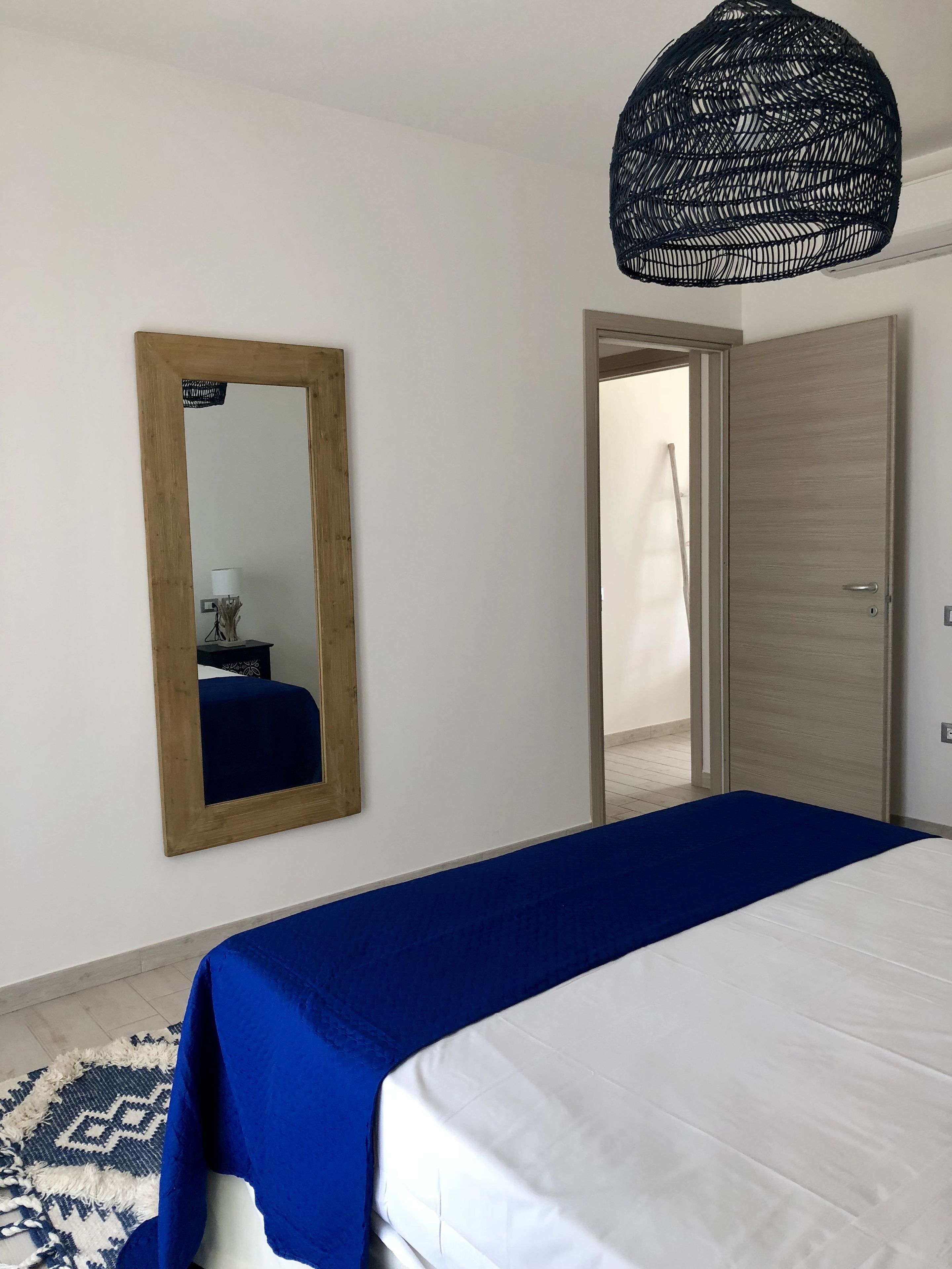 A bedroom with a bed, a wooden-framed mirror, a door, and a hanging light fixture.