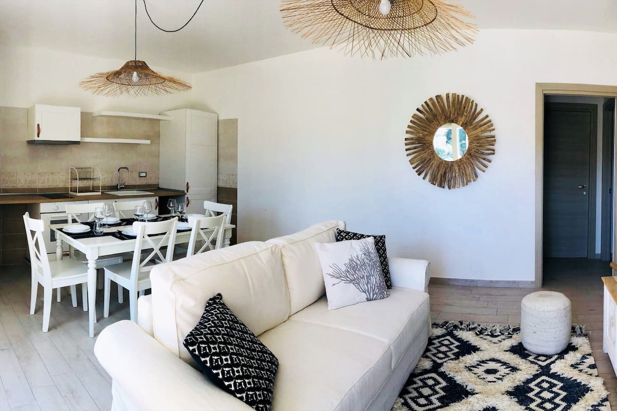 A modern living room with a white sofa, dining table, and chairs. There is a kitchen in the background with white cabinets and a hanging light fixture. A decorative mirror and a television are mounted on the wall.