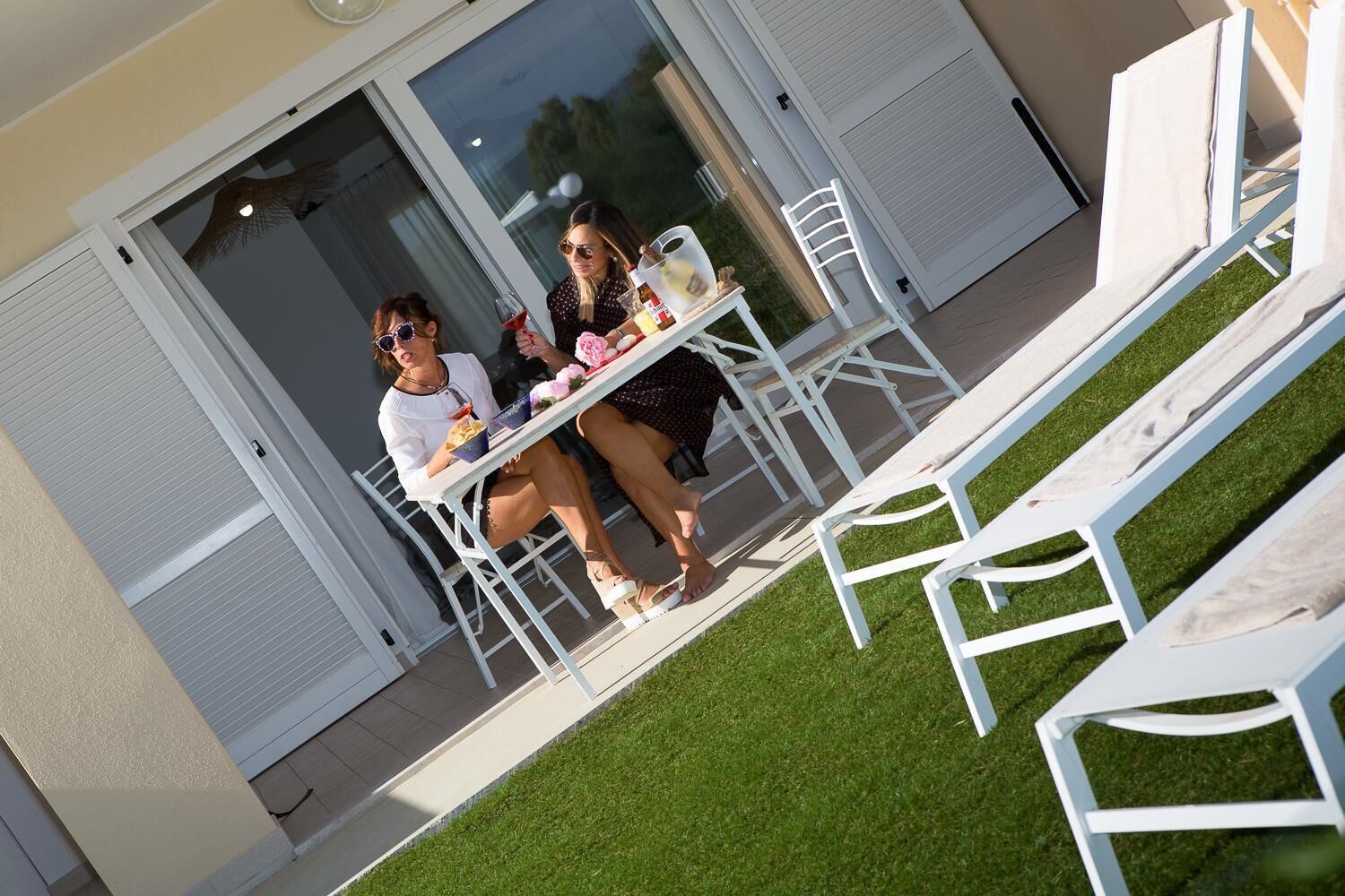 An outdoor patio with white metal chairs and tables, a small lawn, and a building entrance with glass doors.