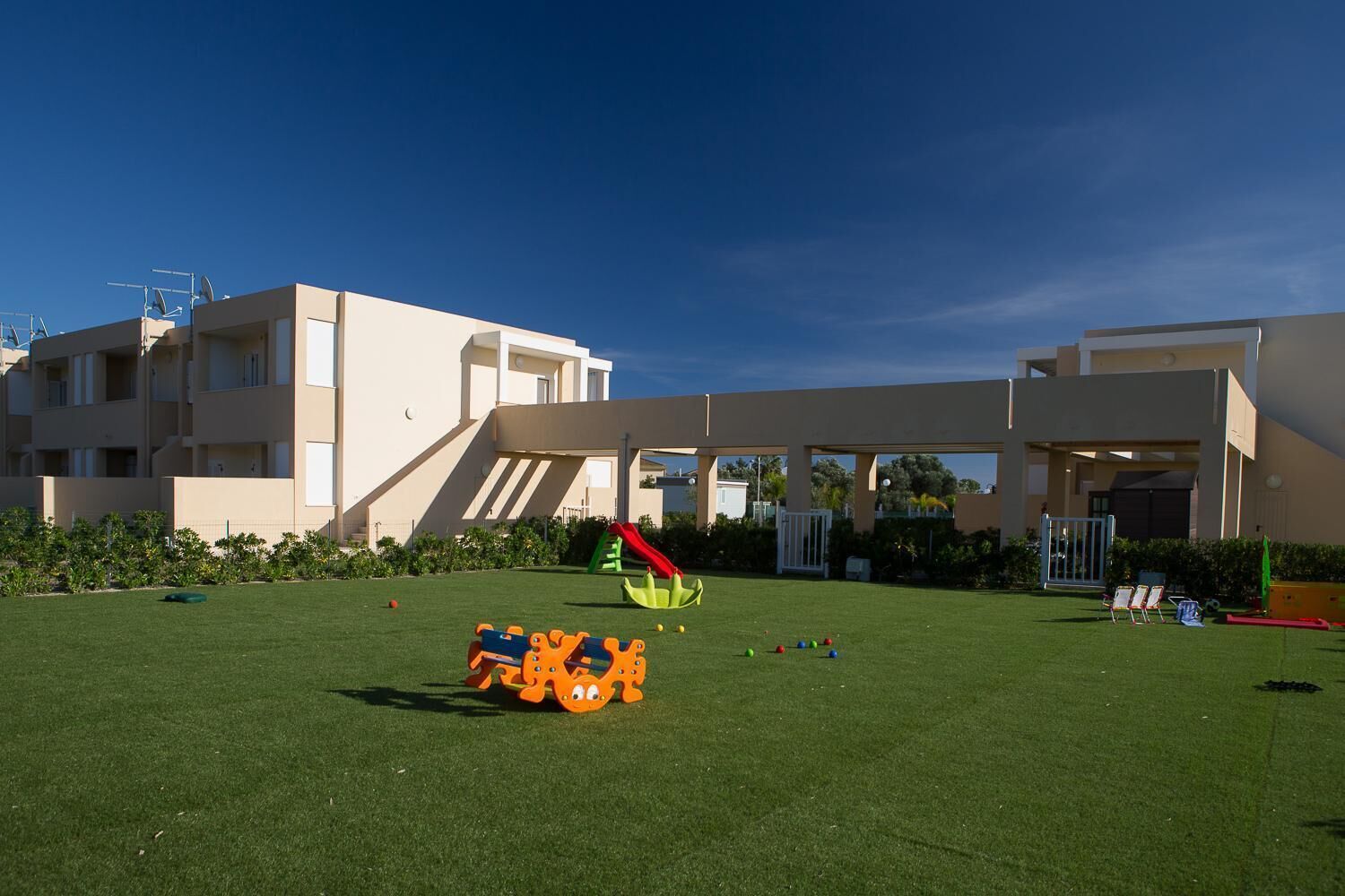 Zona de juegos infantiles con un tobogán y estructura para escalar, rodeada por un edificio con varios balcones y un cielo despejado.