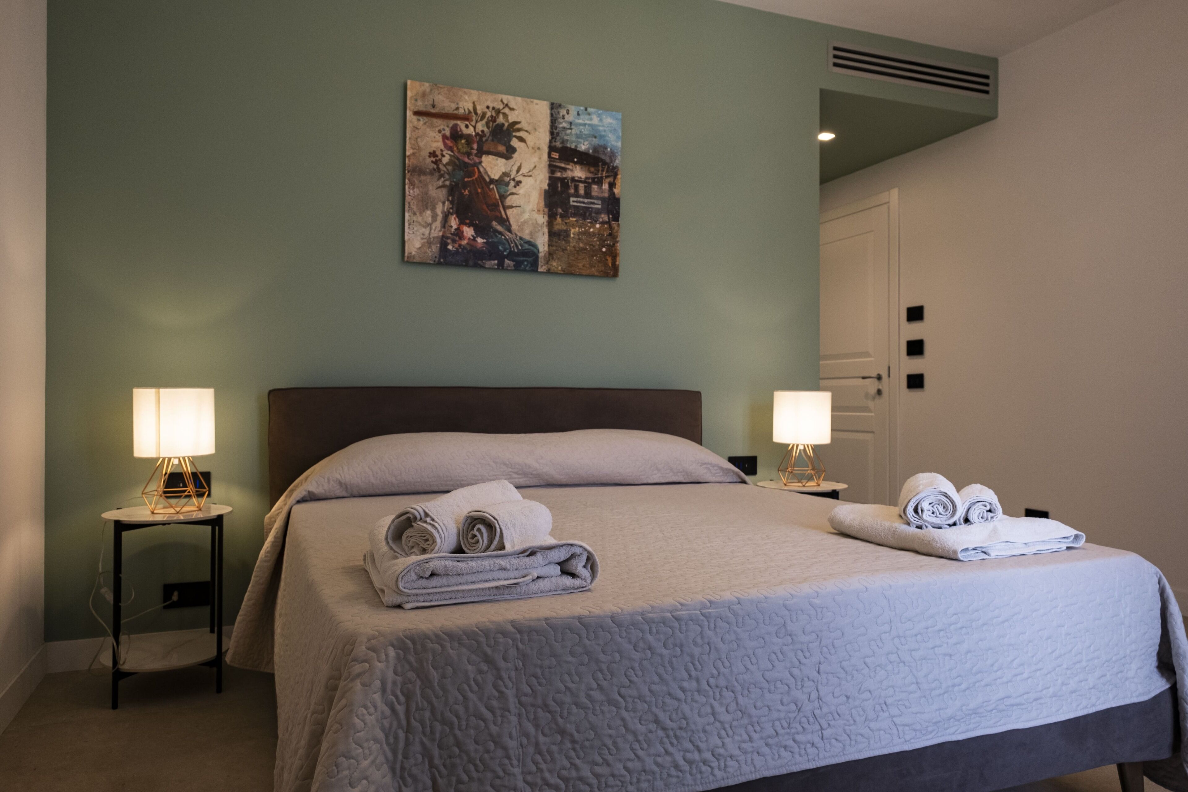 A neatly made bed with folded towels, a wall-mounted painting, and a bedside lamp.