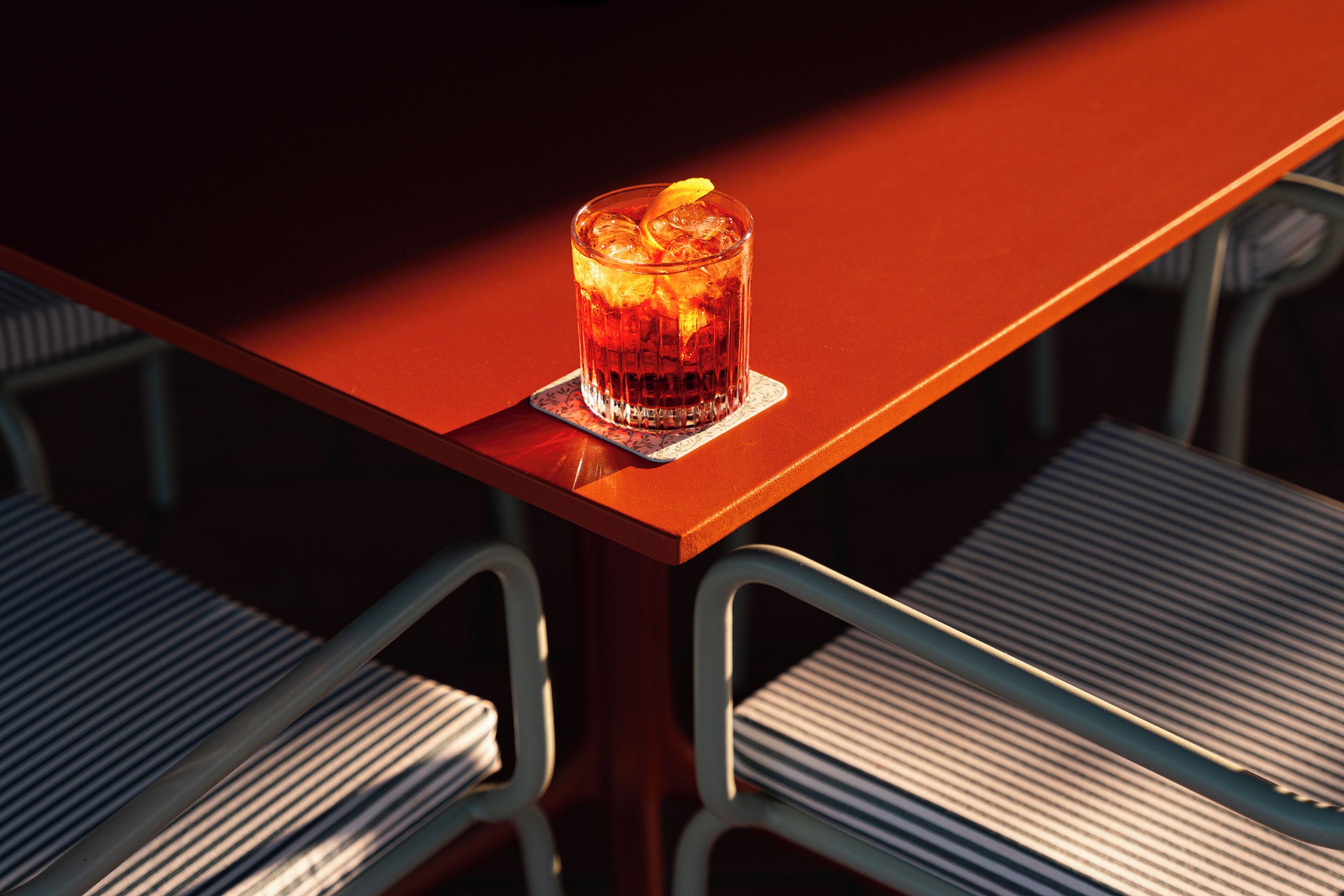 A glass of iced beverage on a coaster, a metal chair, and a wooden table.