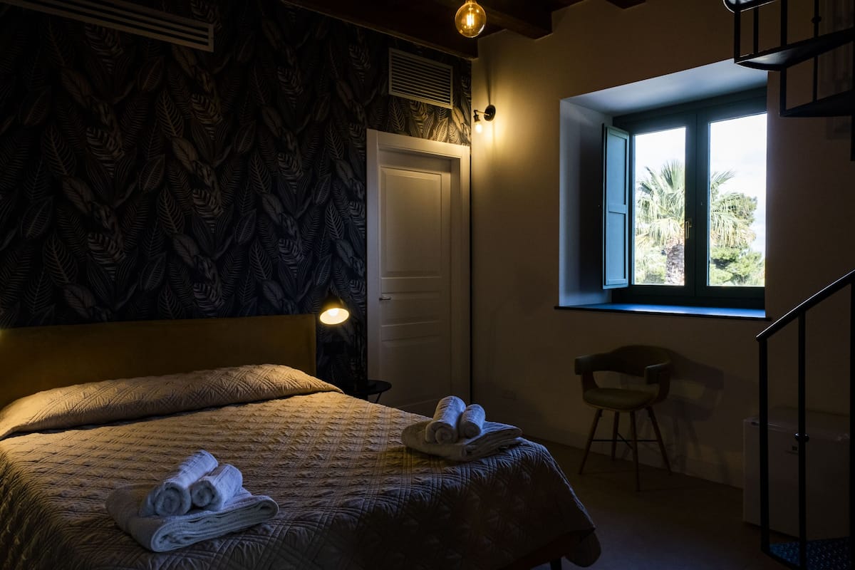 Une chambre à coucher avec un lit, une chaise, une fenêtre donnant sur des palmiers et une porte.
