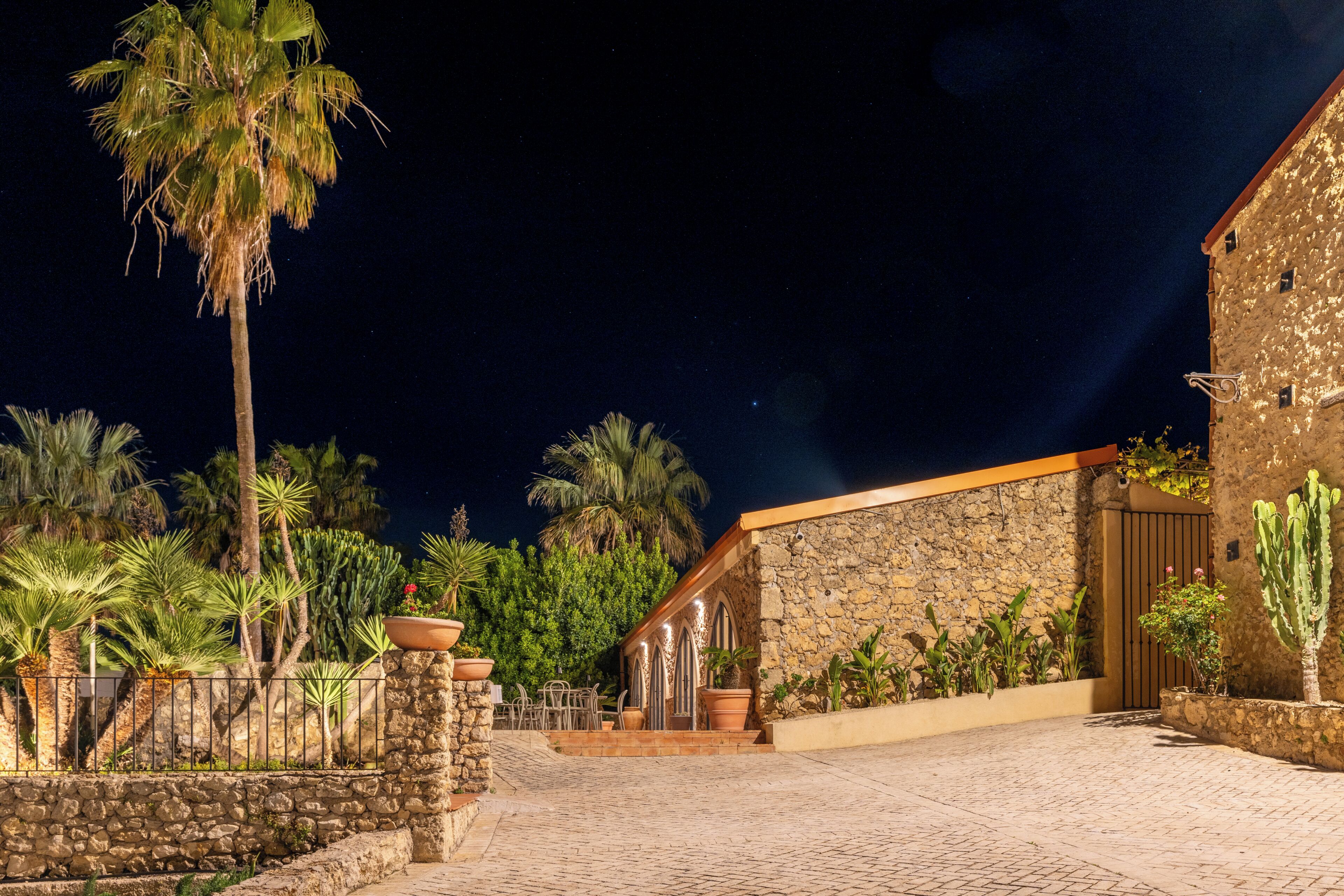 Un edificio de piedra con una entrada porticada, un patio empedrado y varias plantas, incluyendo palmeras y cactus.