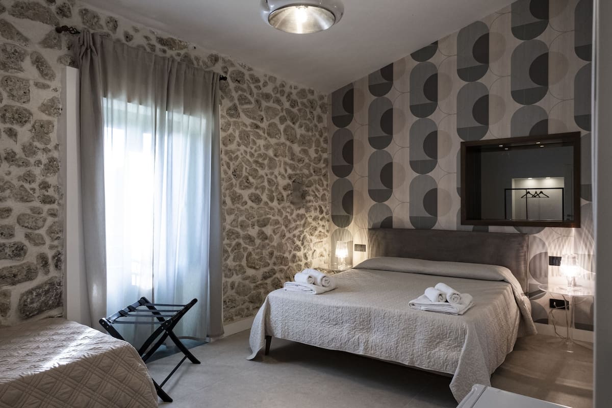 Une chambre avec un mur en pierre, un lit recouvert d’une courtepointe, une table de chevet avec une lampe et un téléviseur fixé au mur.