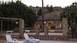 Un área de piscina con dos sillas reclinables blancas, un edificio de piedra con ventanas arqueadas y una planta de cactus.