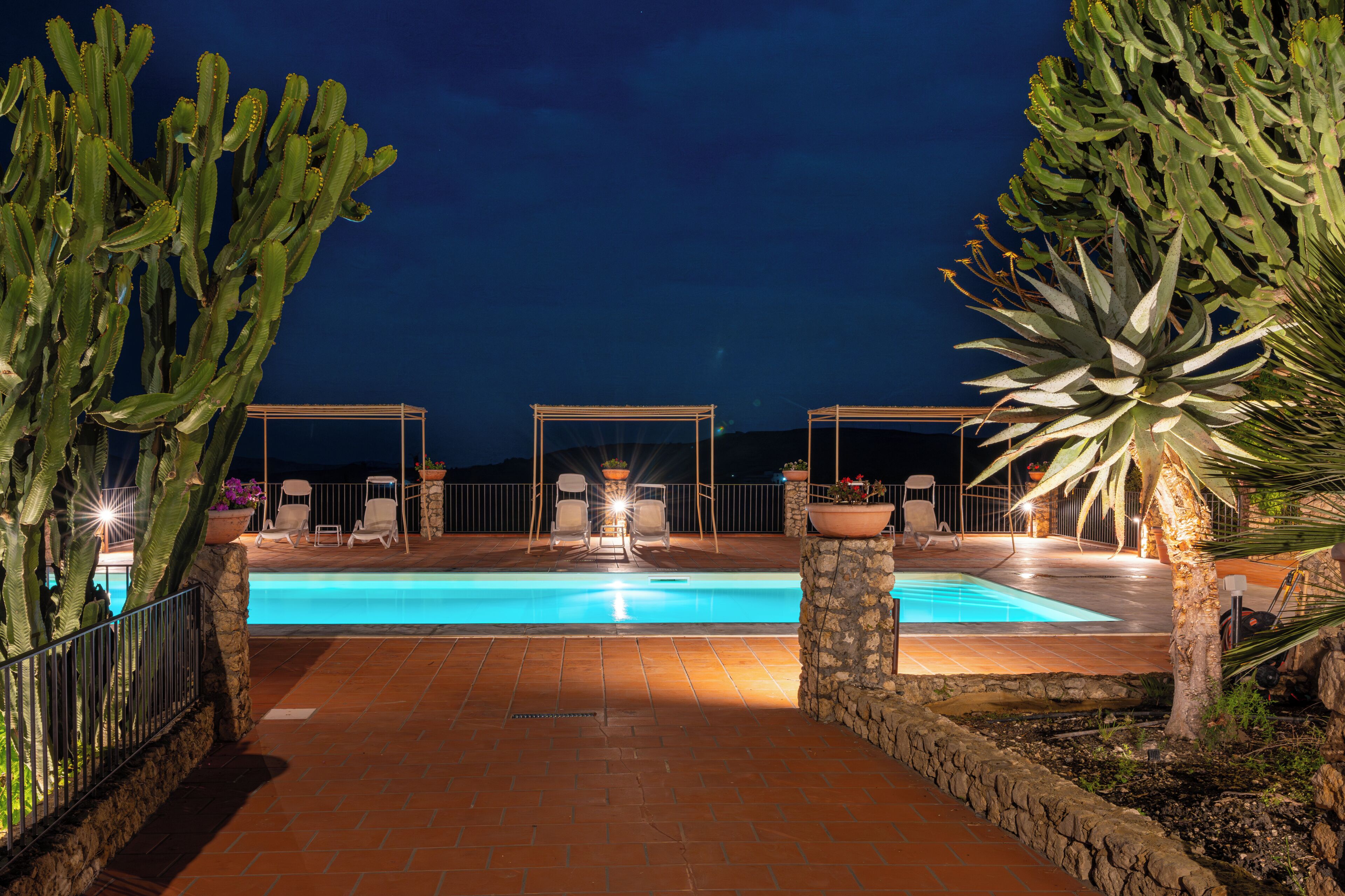 A nighttime pool area with chairs and tables under pergolas, surrounded by cacti.