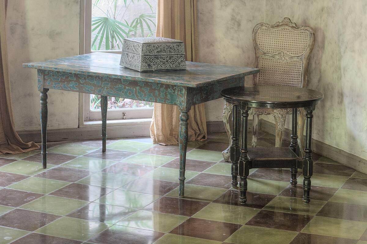 A room with a checkered floor, a table with a decorative box, a chair, a window with curtains, and a hanging light fixture.