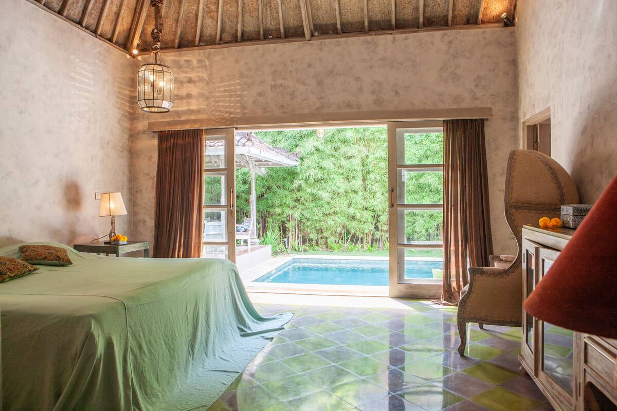 A bedroom with a bed, a nightstand, a lamp, a chair, a vanity, and a view of a swimming pool and greenery outside.