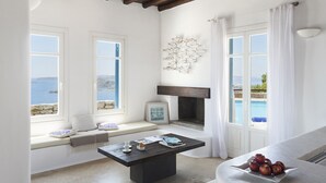 A room with a stone bench by a window overlooking a pool, a fireplace, and a table with apples. - AGL Luxury Villas (Mykonos)
