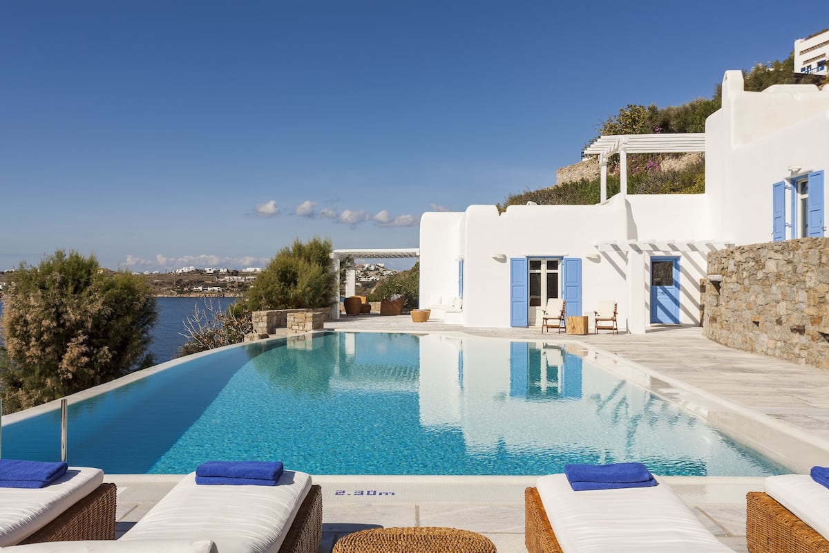 A pool with lounge chairs, a stone wall, and a view of the sea.
