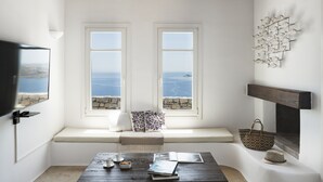 A room with a large window overlooking the sea, a fireplace, and a wooden table with books and a basket. - AGL Luxury Villas (Mykonos)