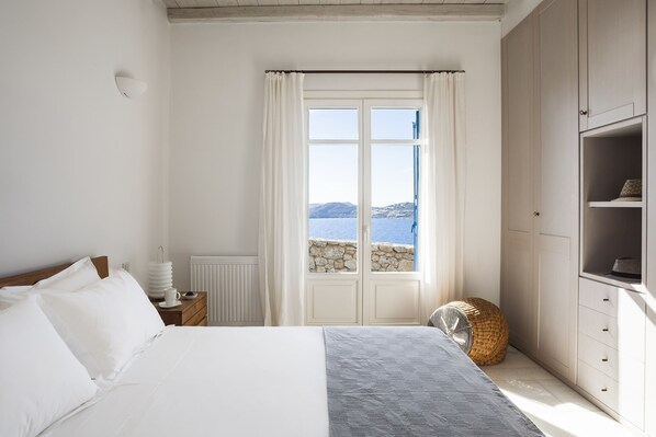 Un dormitorio con cama, mesita de noche, silla de mimbre, armario y ventana con vista al mar.