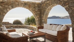An outdoor seating area with stone arches, a wicker sofa, and a table with books and a glass. - AGL Luxury Villas (Mykonos)