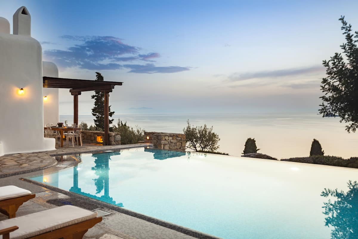 A serene infinity pool with a stone structure and lounge chairs, surrounded by a white building and a view of the sea.