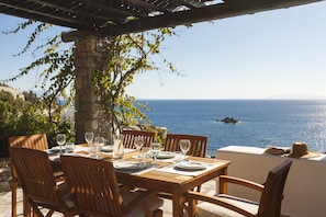A wooden table set for dining with a view of the sea and a stone pillar. - AGL Luxury Villas (Mykonos)