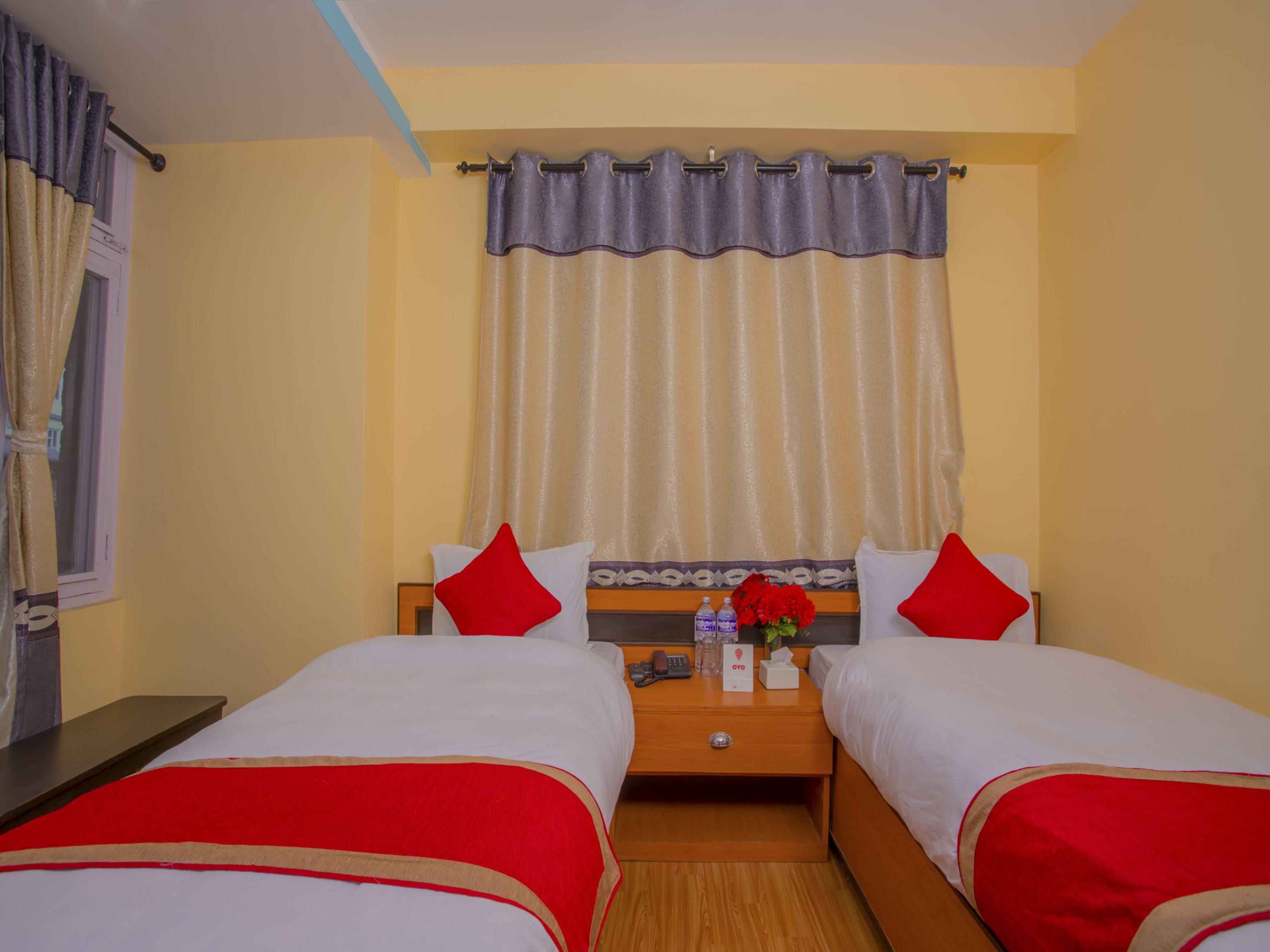 A hotel room with two beds, a wooden nightstand, a telephone, a vase with flowers, and a curtain.