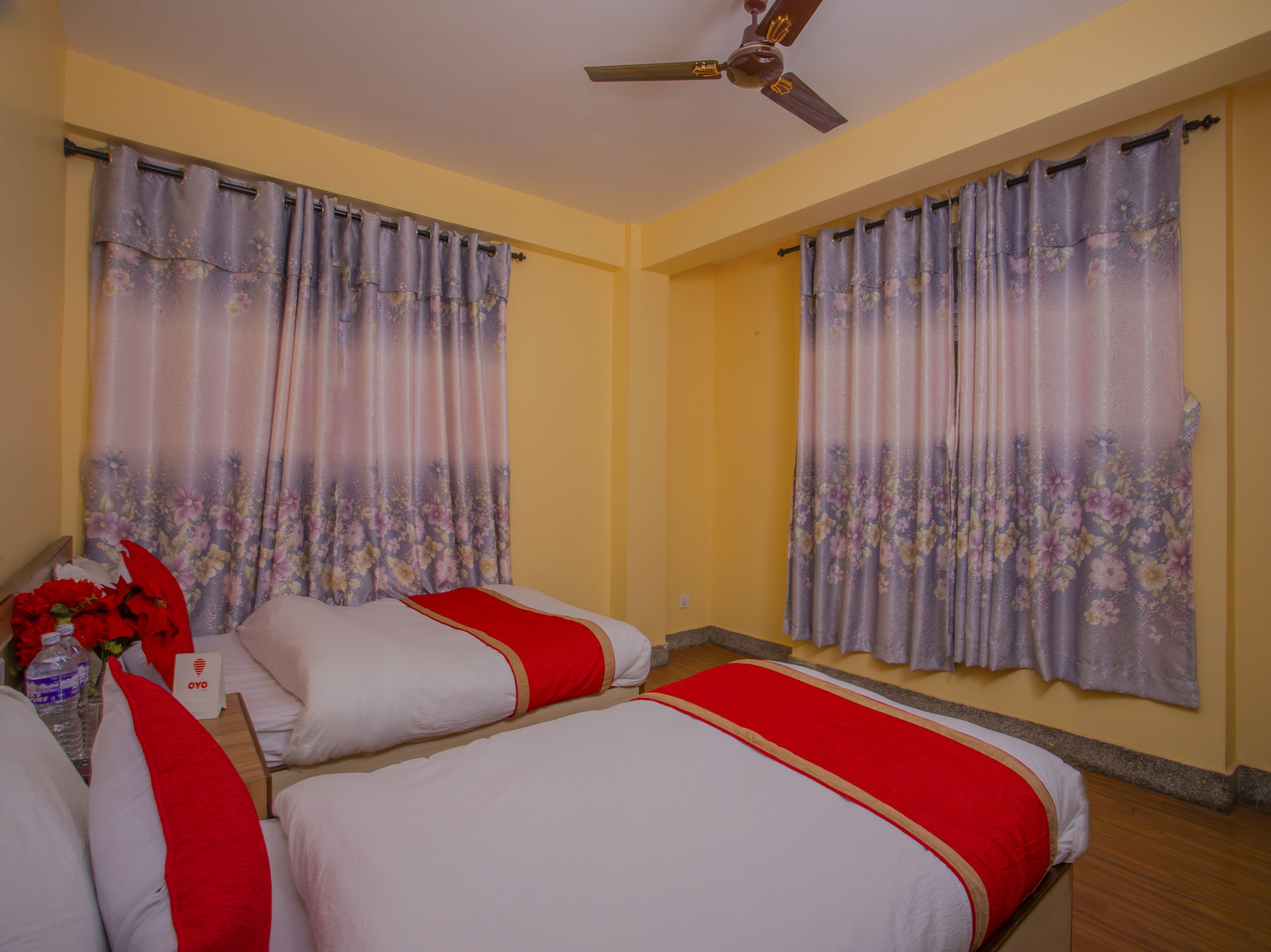 A hotel room with two beds, floral curtains, a ceiling fan, and a small table with a vase of flowers.
