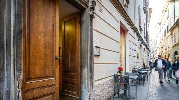 A narrow street with a wooden door and a sign indicating no smoking. There are outdoor tables and chairs, and a few people walking by. - La Contrada del Pittore (Como)